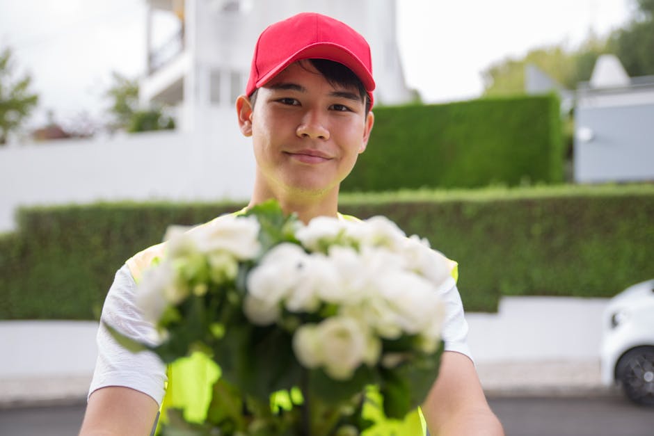 A delivery person wearing a red cap and a reflective yellow safety vest is holding a bouquet of fresh white flowers, possibly roses or lilies, wrapped in clear cellophane. The person's face is smiling