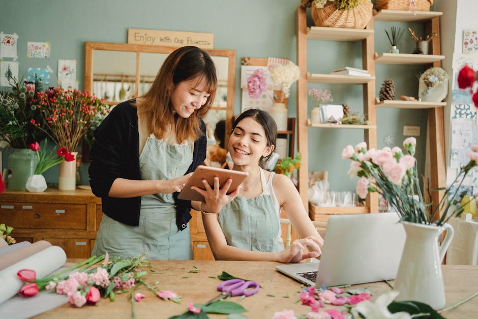 Two women working together in a flower shop, one standing and holding a tablet, the other seated at a wooden worktable. The seated woman has dark hair and is smiling as she looks at the tablet. The st