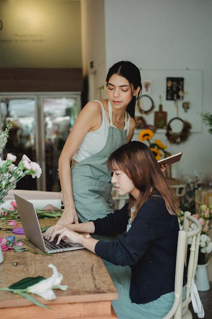 Two women in a flower shop interior are engaged in arranging and checking floral arrangements on a wooden table. The woman standing has dark hair tied back and is wearing a light grey apron over a whi