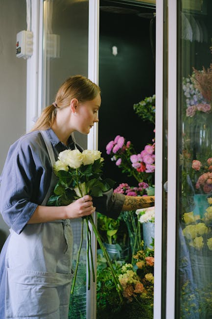A young woman with light brown hair tied back in a ponytail is selecting a bouquet of white roses with rich green foliage from a floral display at Woodside Green florist. She is dressed in a grey apro