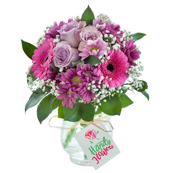 This vibrant, thoughtfully curated bouquet embodies the spirit of Woodside floristry, making it a memorable centerpiece for celebrations or everyday moments. Several pale lavender roses bloom at the core, each layer of petals curling gracefully outward, set off by the luminous presence of hot pink Gerbera daisies whose dark centers draw the gaze. Adorning the ensemble, rich magenta and deep plum chrysanthemums offer a striking depth and complexity, their fine, radiating petals catching subtle shifts of daylight, reminiscent of the artistry seen around Woodside's nearby community centers. Interspersed between the bold hues, baby's breath-delicate and snow-white-softens the palette and adds a diaphanous quality to the compact arrangement. Contrasting the floral profusion, lush dark green leaves peek out from beneath and between the blossoms, echoing the greenery of Woodside's neighborhood parks. The bouquet rests in a clear glass vase with a unique crackled texture, its surface glimmering with reflections, rooted firmly yet elegantly on a tabletop. Around the neck, a strand of natural raffia is tied, its rustic charm evoking a handmade touch, with a crisp white gift tag attached-ready for a handwritten sentiment. This composition, ideal for speedy delivery to homes and offices around Woodside, brings together local expertise, vibrant color, and the emotional resonance of a truly personal floral creation, perfect for birthdays, anniversaries, or simply to share a moment of beauty with someone special.